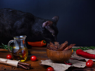Black cat smelling meat jerky in bowl on wooden table in front of black background. Olive oil in glass jug, meat sticks, red pepper, cherry tomatoes and rosemary on rustic craft paper and brown wood