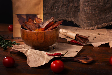 Jerky snacks in wooden bowl, olive oil in glass jug, meat sticks, red pepper, cherry tomatoes and rosemary on rustic craft paper and brown wood in front of burlap cloth background. Dried meat.
