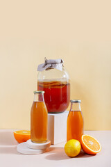 Glass gar with komucha tea, citrus fruits and two bottles with ready beverage on modern podiums in front of yellow background with copy space. Orange and lemon. Healthy fermented drink. Vertical