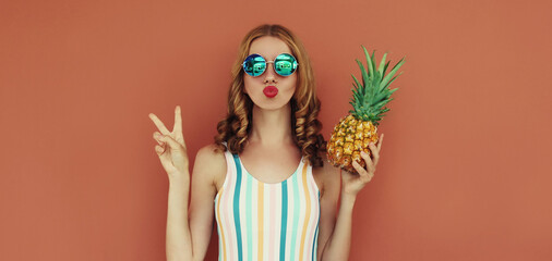 Summer portrait young woman blowing her lips with pineapple wearing a sunglasses posing on a brown background