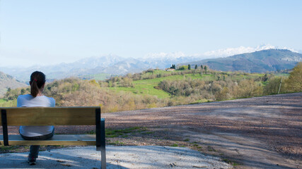 Young woman seen from her back sitting alone on a wooden bench, looking to a beautiful green landscape on a sunny day. Picos de Europa mountains with snow in the background.