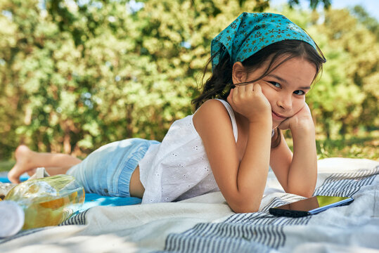 Horizontal image of a little girl lying on the blanket playing on the device in the park. Kid having cozy summer picnic with family with fresh lemonade and fruits outdoor.