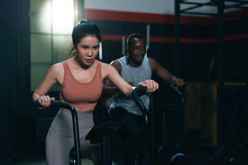 Multi ethnic group of young athletes training on stationary bike cardio machine building muscle in fitness gym.