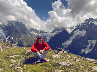 Naklejka na ściany i meble Young man sitting on mountain peak in cloudy weather. Adult guy rests, enjoying amazing mountain landscape in summertime.