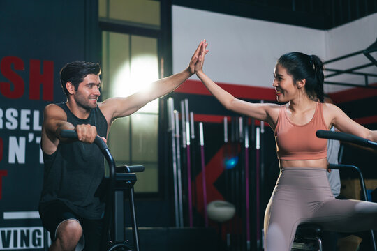 Asian female and Caucasian male athletes or sport people giving high five together after successed training on stationary bicycle. Health care and fitness in gym concept
