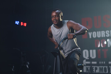 Fototapeta na wymiar African ethnicity man listening music while using spinning bike on cardio machine in fitness gym