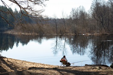 A lone fisherman sits on the shore in a spring reservoir and catches fish with a fishing rod