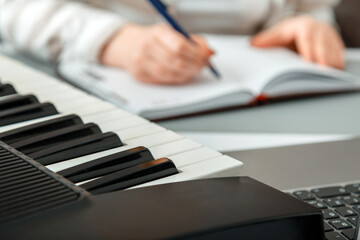Woman composes music, female hands write notes or song in music book. Teen girl learns to play piano music, makes notes in notebook. Synthesizer piano keyboard instrument. Online music education