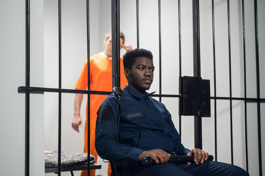 A black prison guard sits in front of cages with dangerous criminals on a chair with a baton.
