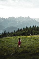 Fototapeta premium Back view of slim young woman walking on grassy valley on background of green mountains on cloudy evening