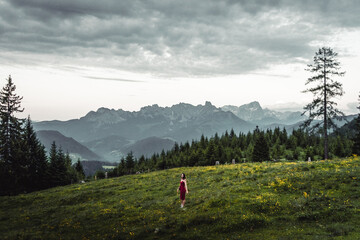 Naklejka premium Back view of slim young woman walking on grassy valley on background of green mountains on cloudy evening