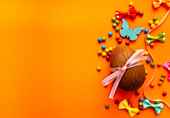 Chocolate Easter egg with pink ribbon, candy, bow and butterfly on orange background. Spring holidays concept