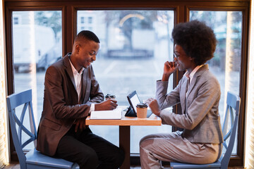 Two african business partners sitting at cozy cafe during working meeting. Young man and woman using modern devices indoors. Coworking process.