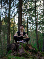 Naklejka na ściany i meble A young beautiful tanned teenage girl is doing yoga in a green forest with tall trees, in nature. Eyes closed. The lotus position. Black sportswear. The concept of a healthy lifestyle.