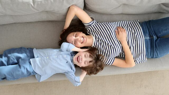 Top above view of happy young mom and little kid son relaxing on sofa at home. Couple of carefree mother and cute child lying, resting on couch, having fun, looking together at camera. Family, love.