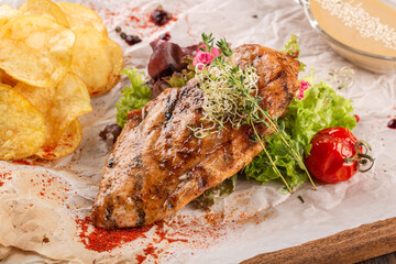 Grilled chicken fillet on a board garnished with tomatoes, herbs, chips and sauce.