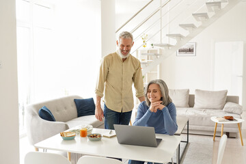 Happy middle aged mature 50s family couple having fun using laptop computer at home. Smiling senior older mature husband and wife laughing watching tv online or having virtual meeting in living room.