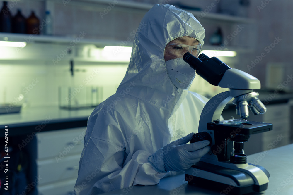 Sticker Female chemist in protective workwear and in mask looking through the microscope while working at the table in the lab