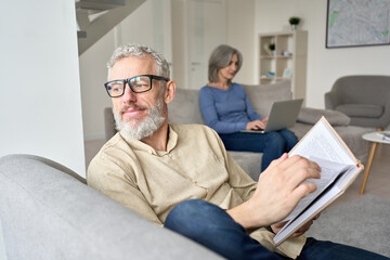 Happy older senior man husband wearing glasses reading book relaxing sitting on couch at home with mature wife using laptop. Middle aged family couple enjoying casual daily activities