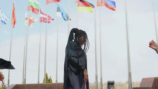 Three young women of different nationalities joyfully throw hats up against many flags of different countries fluttering in the wind on the background. Outdoor view