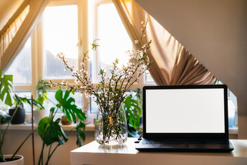 A home workplace with white screen laptop for mockup, blooming branches in a vase on the desk with room interior background in sunset light. Freelance, working from home, online learning, home office - Powered by Adobe