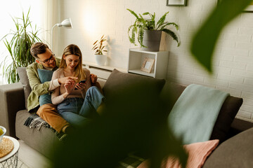 Photo of happy young couple, relaxing on sofa and shopping online using a cellphone.