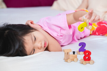 Obraz na płótnie Canvas Asian cute girl lying playing wooden toys on a soft white mattress. Adorable kid play number train. Little child's relax moment. Children aged 4 years old wearing pink shirt on the bed.