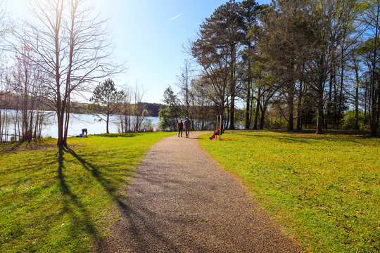 People Walking Down A Hiking Path Near The Lake With A Woman In Red Sitting On A Bench With Lush Green Grass And Trees And Vast Lake Water And Blue Sky At Lake Horton Park In Fayetteville Georgia USA