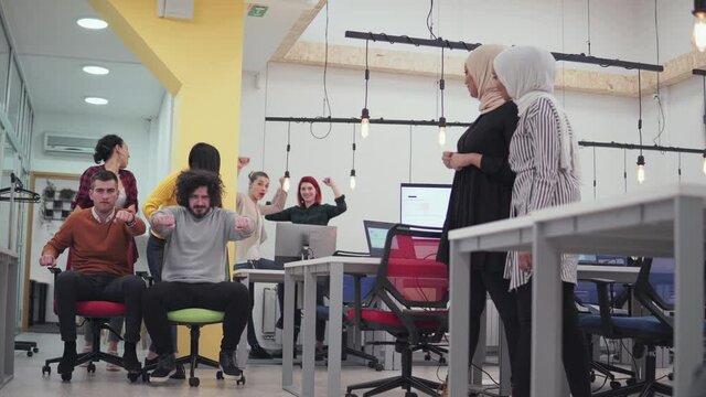 Multiethnics startup business team of software developers having fun while racing on office chairs,excited diverse employees laughing enjoying funny activity at work break
