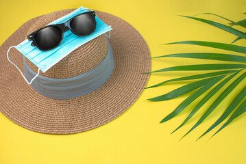 Tropical palm leaves, straw summer hat, sunglasses and blue medical mask on yellow background. Travel safe.
