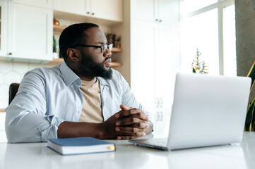 Successful handsome african american bearded man with glasses, freelancer or employee, works at home uses laptop, looks thoughtfully aside, thinks about business project, hands folded in front of him