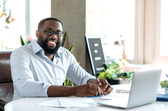 Successful handsome confident bearded african american businessman, real estate agent or ceo dressed in formal stylish clothes and glasses, sits at workplace, looking at camera, smiling friendly