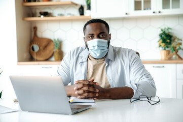 Portrait of a young African American man in a medical mask, in stylish clothes, working remotely, sitting at a table at home, looking at the camera, uses laptop. Distance work during quarantine