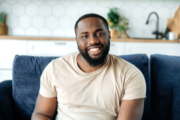 Close-up portrait of handsome happy young confident african american bearded man, sitting on sofa at home in living room, wearing t-shirt, looking at camera and smiling friendly
