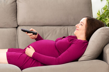 Shot of a pregnant woman watching tv content in the day sitting on a couch in the living room at home