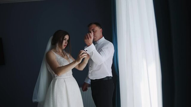 The bride helps the groom get dressed and prepare for the wedding ceremony. A young couple stands by the window and the wife fastens her husband's cufflinks on the sleeves of his shirt. Care