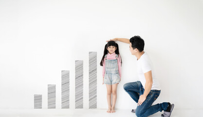 Dad measures height increase of her child daughter at white brick wall with graph growing growth
