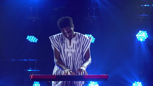 African American man plays piano synthesizer keyboard and sings in dark studio with blue lights. Musician with white ethnic pattern on his face in national striped shirt performs on stage.