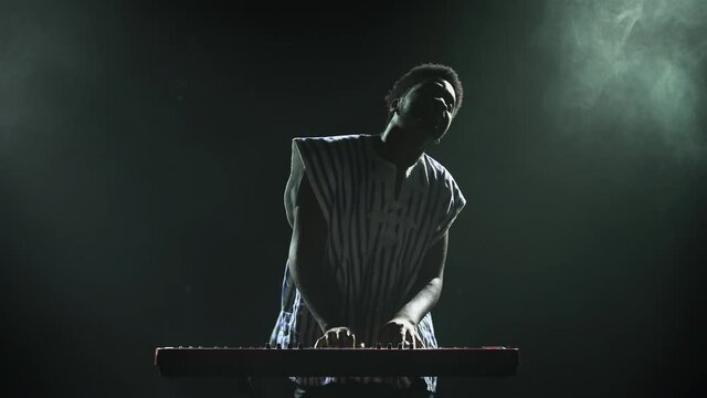 Silhouette of african american man playing piano synthesizer on stage in dark smoky studio. Black musician with a white ethnic pattern on his face performs against the background of lights.