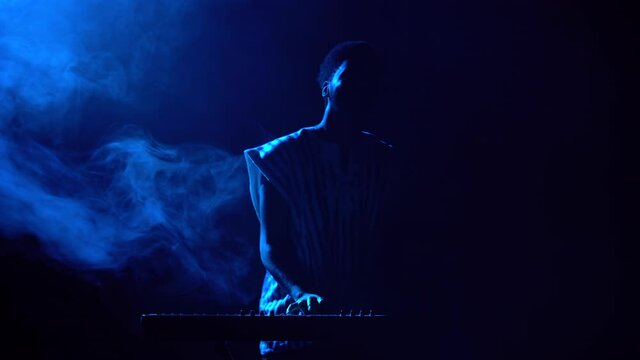 Silhouette of african american man playing piano synthesizer on stage in dark smoky studio. Black musician with white ethnic pattern on his face performs against background of blue lights.