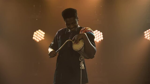 Black musician plays talking African yuka drum in a dark studio against a backdrop of bright lights and smoke. An African American man hits the drum and enjoys the music. African National Folklore.