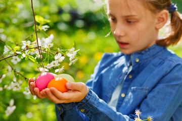 Child on Easter egg hunt in blooming garden with spring flowers. Kid with colored eggs. Little girl picking flower. Easter decoration, family celebration, Christian traditions.