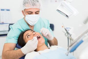 The dentist in face mask inspects female patient teeth with mirror and probe