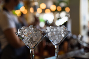 Cocktail glasses filled with ice bokeh partylights in the background with an unrecognisable masked attendant in the background 