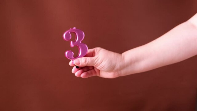 bright pink number three in a female hand, birthday candle on a red background, concept of progress, three years, anniversary