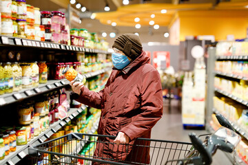 Old senior woman wear medical mask, protection against pandemic coronavirus disease. 90 years retired female lady push cart trolley for shopping in supermarket. Woman buy food and groceries