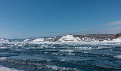 Spring. The ice on the river is cracked, melting snow is gathering between the ice floes. Snow-covered mountains against the background of a blue sky. Ice drift on the Angara.