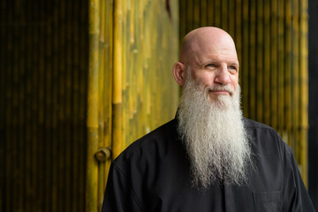 Portrait of man bald man with long gray beard outdoors against bamboo wall thinking