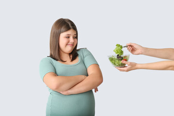 Offering of healthy vegetable salad to displeased overweight girl on light background