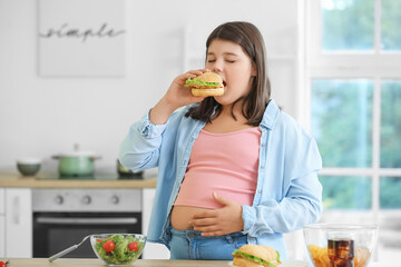 Overweight girl eating unhealthy burger in kitchen